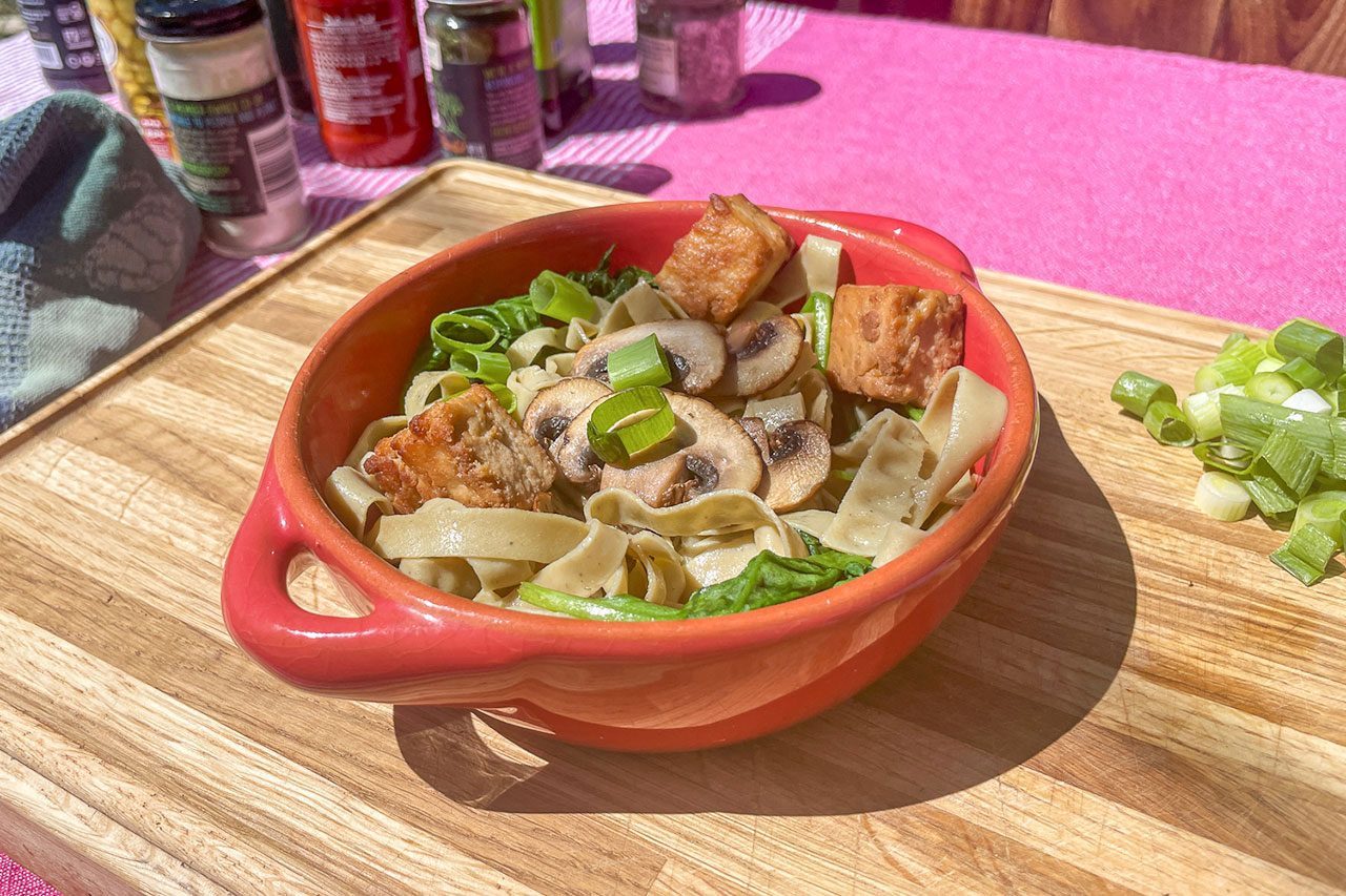A red bowl of pasta with mushrooms, green onions, and tofu sits on a wooden board. Chopped green onions and spice jars are in the background.