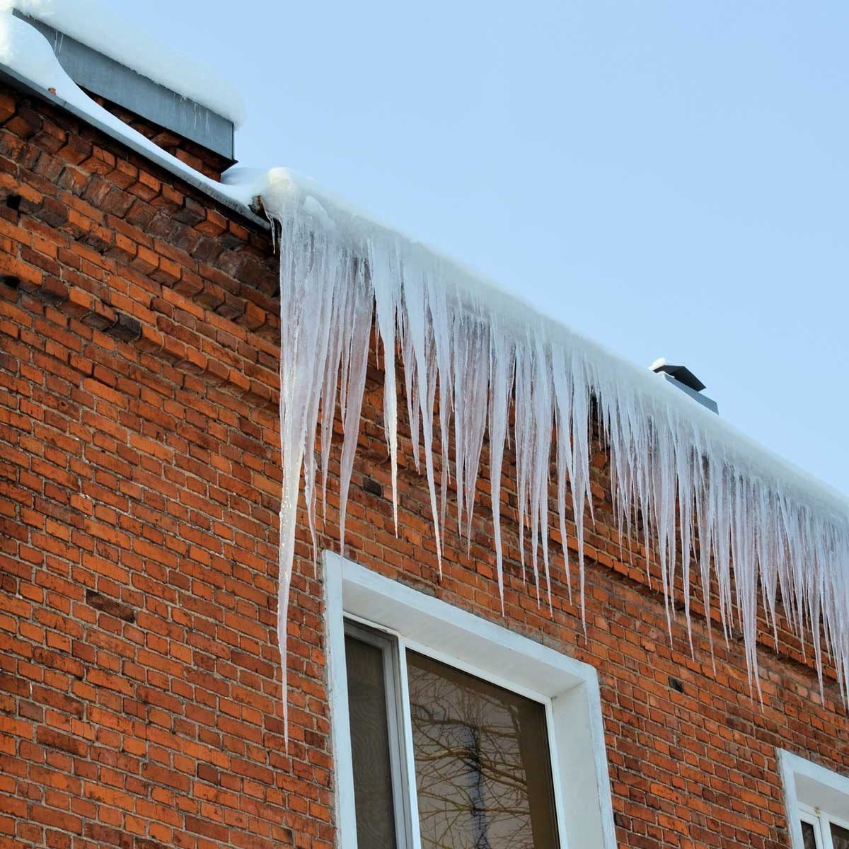 Icicles hang from a snow-covered roof, reflecting sunlight against a backdrop of a brick wall and a window, under a clear blue sky.