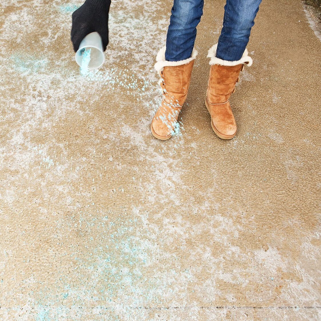 A person in brown boots pours blue granules onto a light-colored surface, preparing to treat a snowy or icy area.