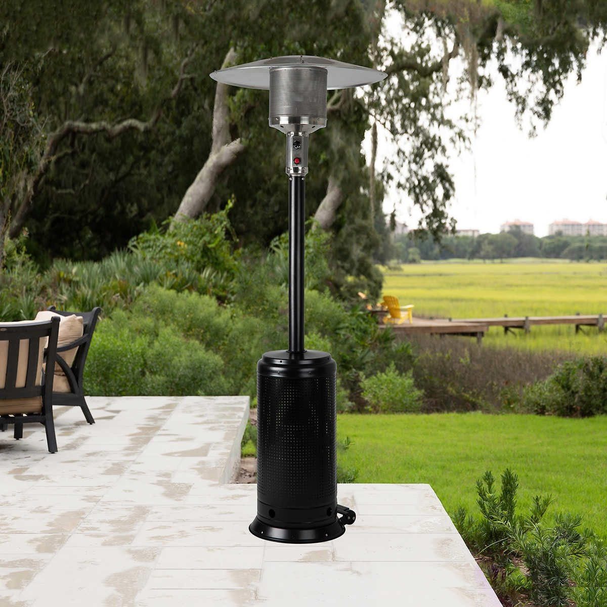 A patio heater stands on a stone floor, providing warmth in an outdoor setting surrounded by greenery and a view of a field and distant buil
