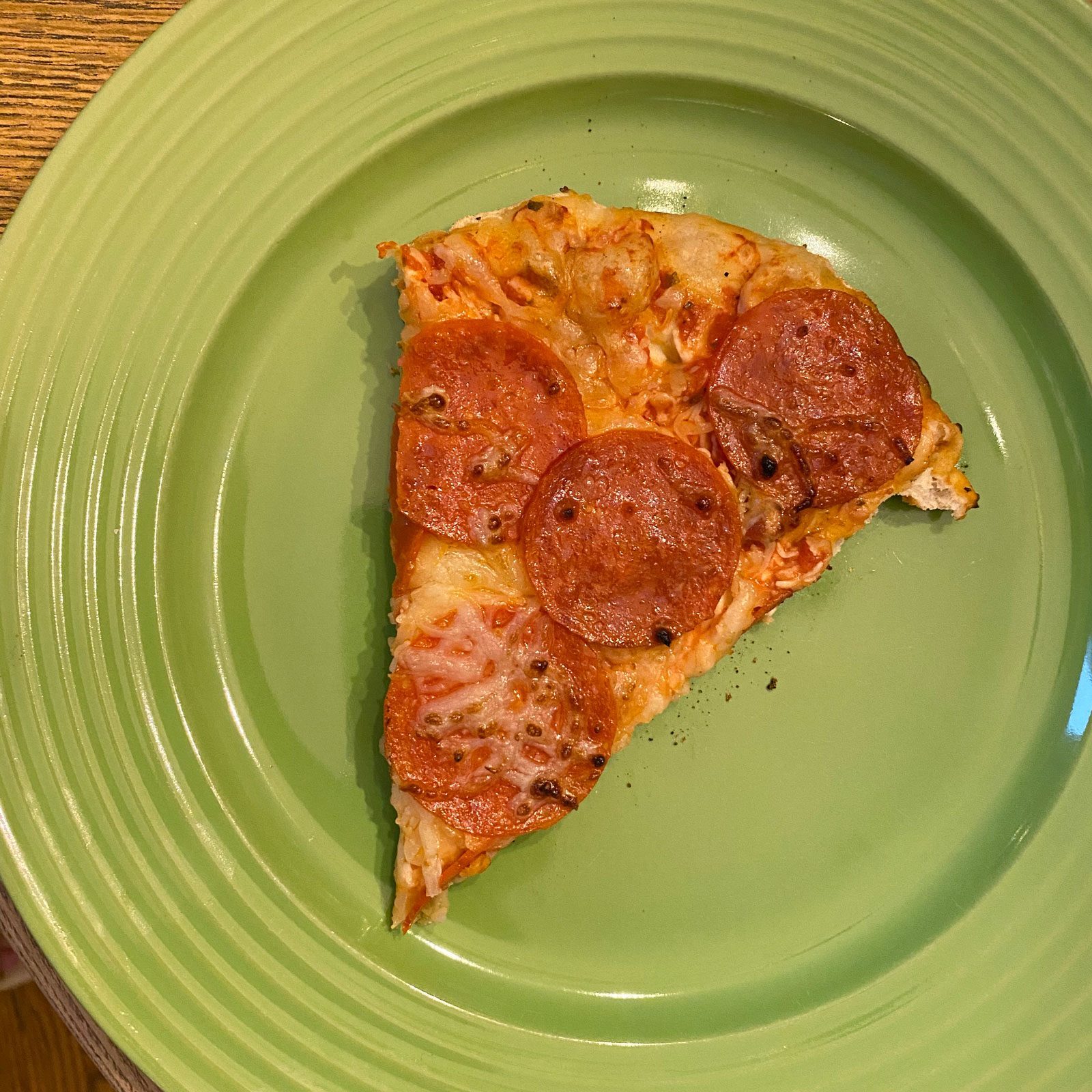 A slice of pepperoni pizza sits on a green plate, with melted cheese and toppings, against a wooden table background.