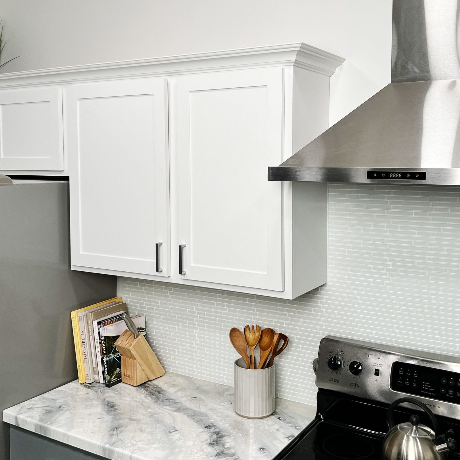 A marble countertop hosts a cup of wooden utensils, cookbooks, and a stainless steel stove, with white cabinets and a sleek range hood above