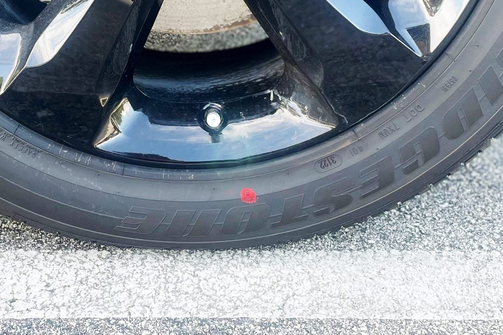 A black car wheel with a shiny rim rests on a paved surface, displaying a red dot on the tire sidewall.