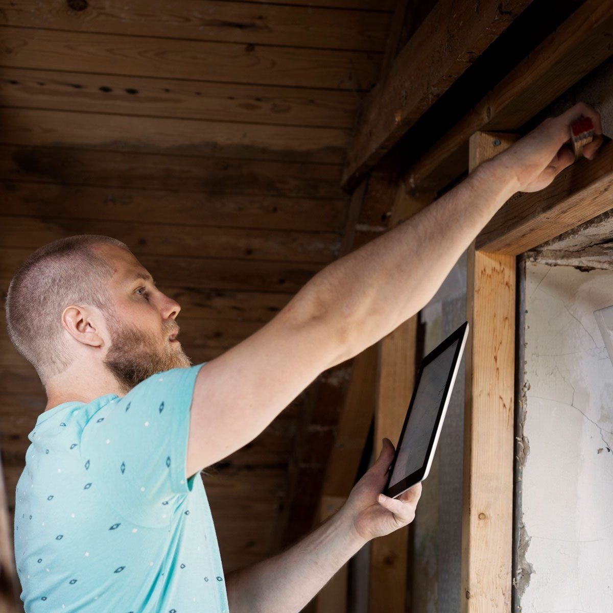 A man with a beard holds a tablet and measures something above a window in a room with wooden beams and a partially finished wall.