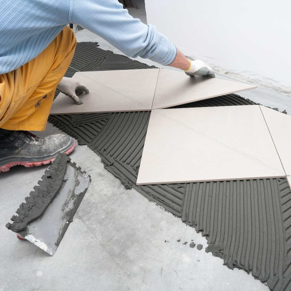 A person places large tiles onto fresh mortar in a room, adjusting their position carefully on a patterned bed of adhesive for flooring.