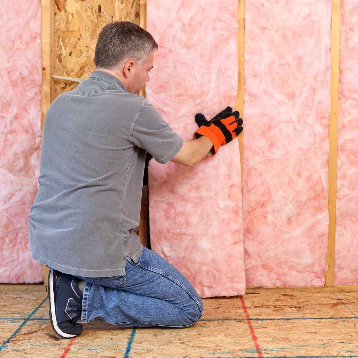 A man kneels, installing pink insulation between wooden framing in a construction environment, wearing gloves for protection while working o