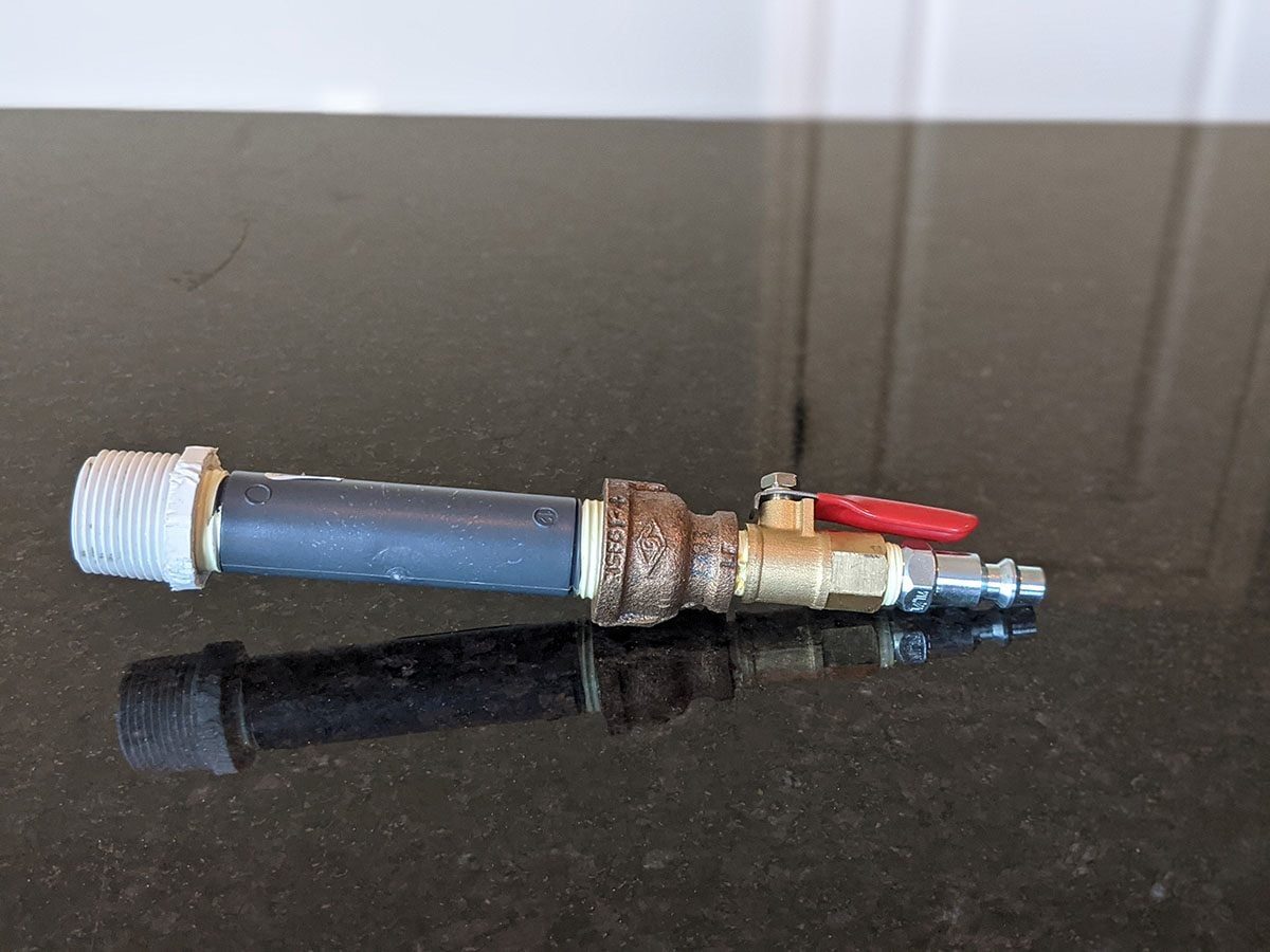 A metal valve with a red lever connects to a gray pipe, resting on a dark, reflective surface in a bright environment.