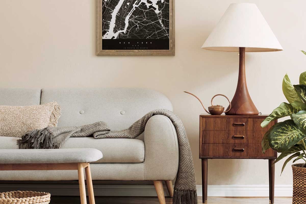A light-colored sofa showcases a cozy blanket and pillows, accompanied by a wooden side table with a lamp, framed map, and a potted plant.