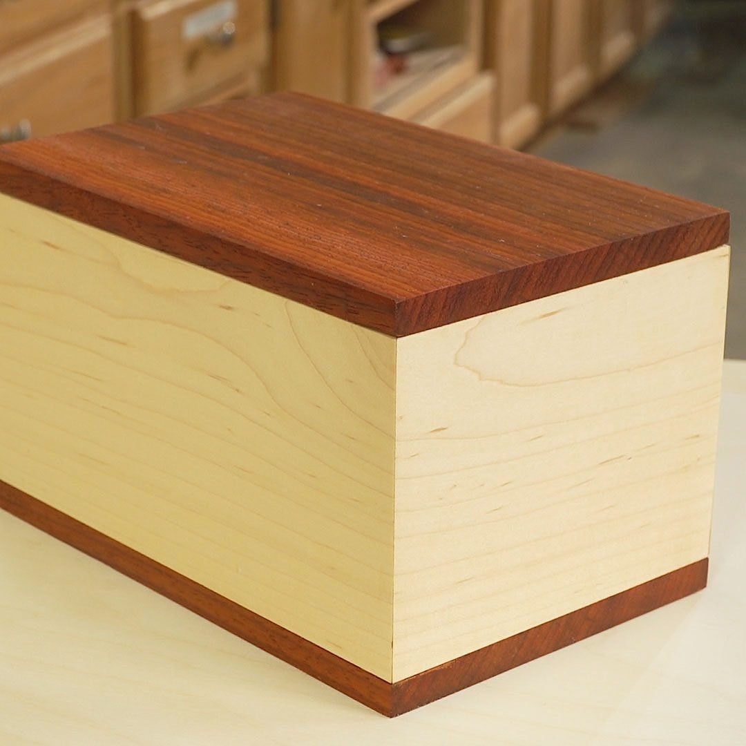 A wooden box with a light-colored body and dark wooden top sits on a surface, surrounded by a workshop environment with cabinets in the back