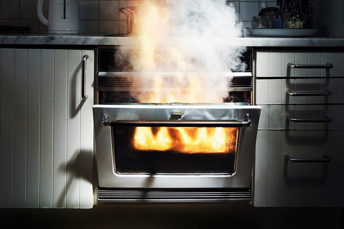An oven emits flames and smoke, indicating a fire. The kitchen's cabinets and countertop are visible, suggesting a domestic cooking environm