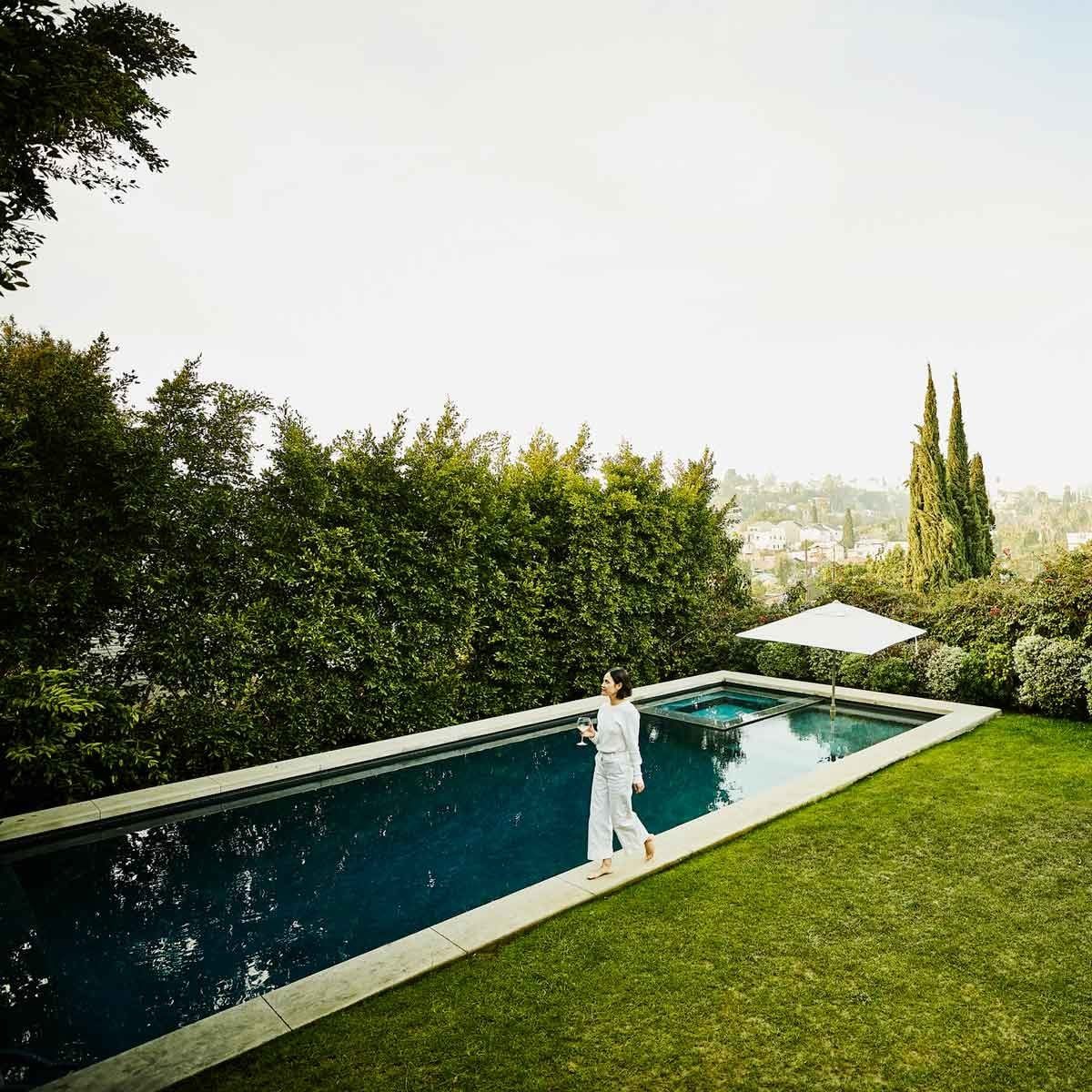A woman walks barefoot alongside a sleek swimming pool, holding a drink, with lush greenery and distant hills in the background under a clea
