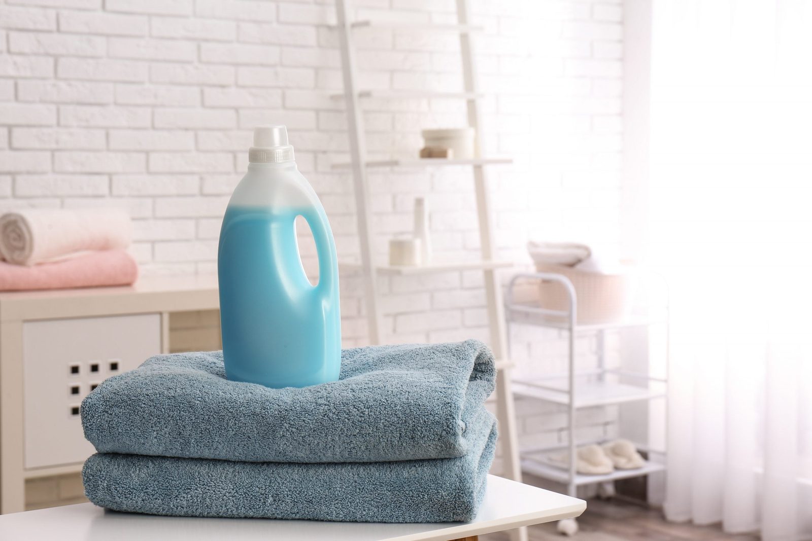 A blue detergent bottle rests on two stacked blue towels in a bright, airy room with white brick walls and soft textiles in the background.