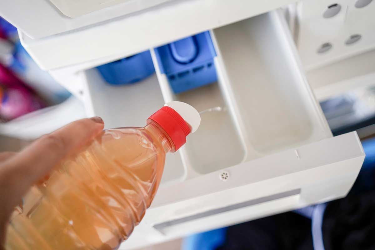 A hand holds a bottle with a red cap, pouring liquid into an empty compartment inside a white appliance, with colorful items visible in the