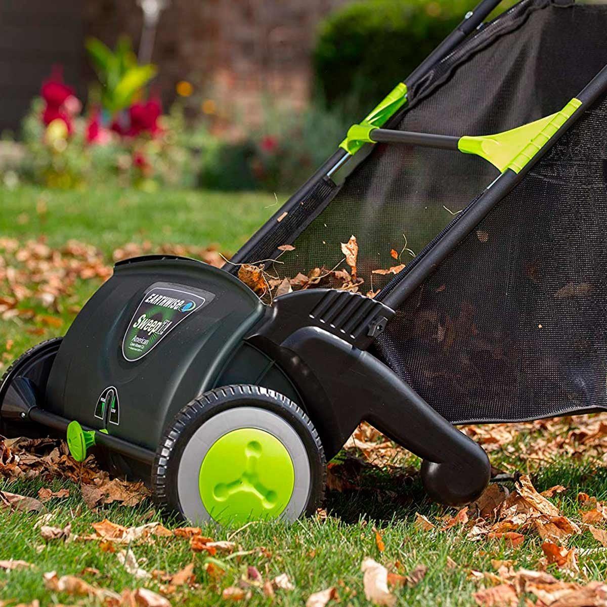 A lawn vacuum collects leaves, moving across a grass surface. The surrounding area includes green plants and colorful flowers in the backgro