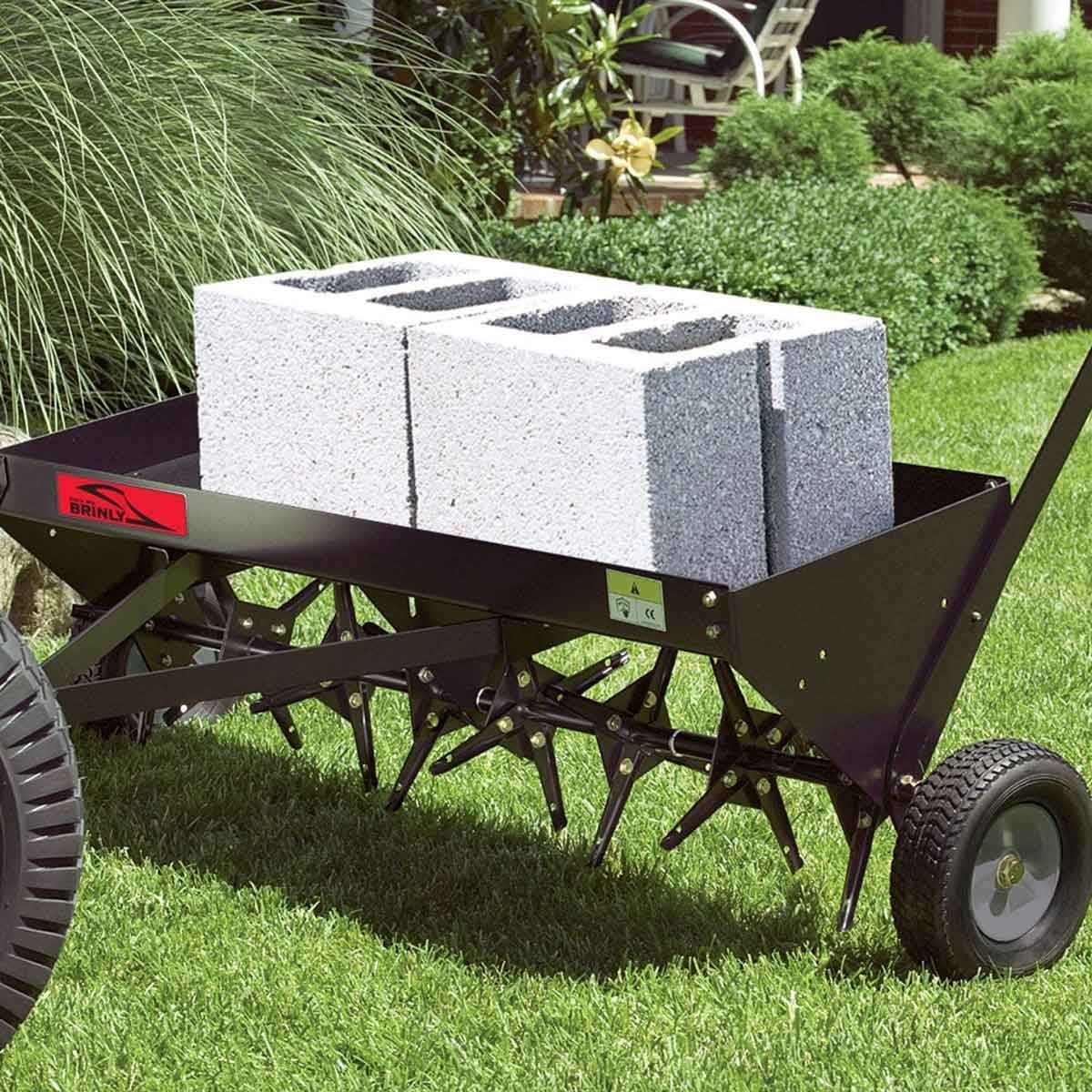A garden tool cart carries three gray concrete blocks on a grassy lawn, with metal tines visible underneath, surrounded by shrubs and a dist