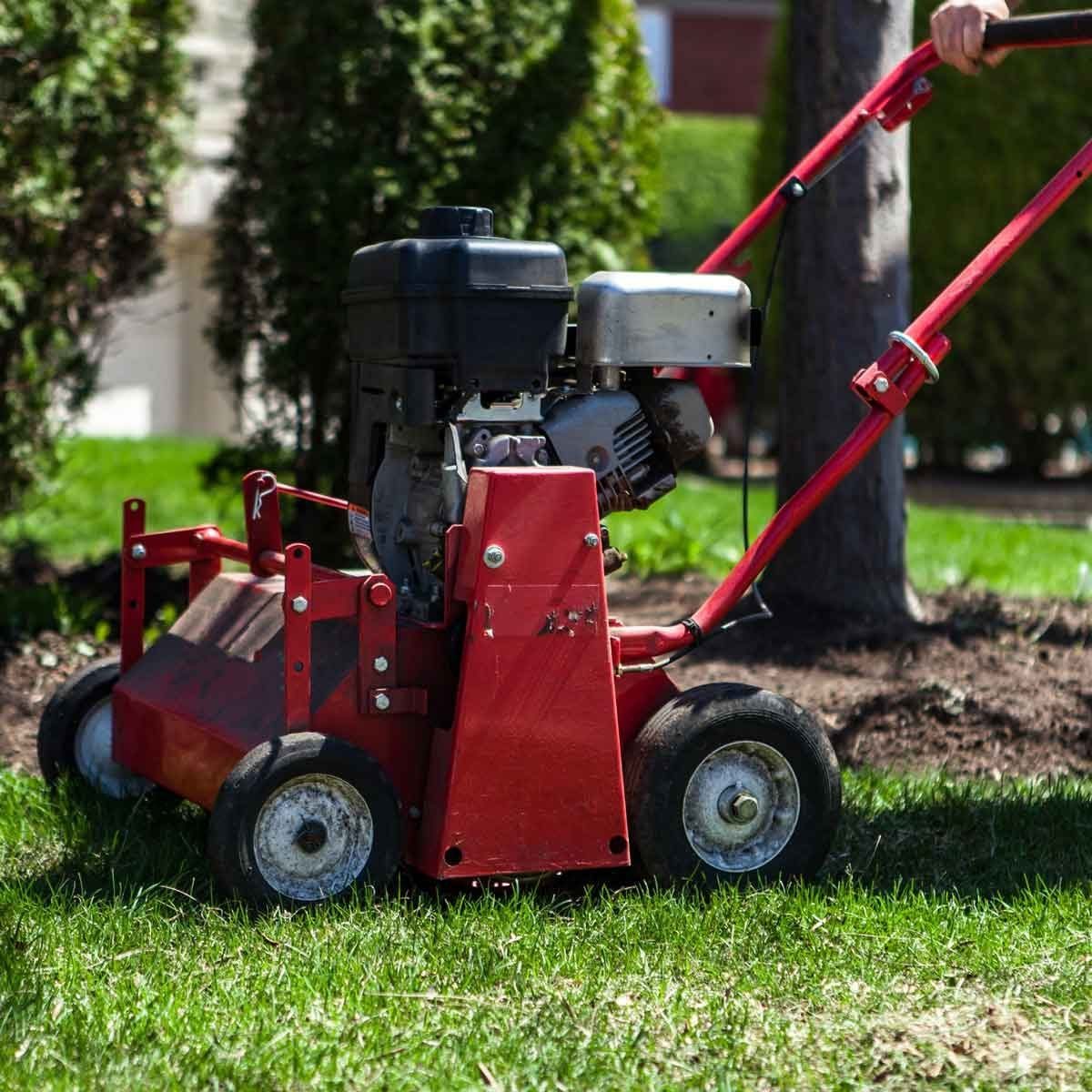 A red lawn care machine moves across a grassy area, preparing the soil for planting, with trees and a house visible in the background.