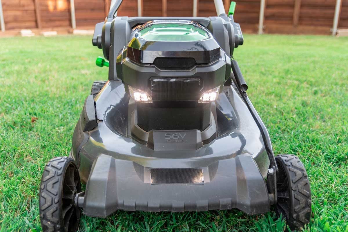 A black electric lawn mower stands on green grass, equipped with a battery compartment and headlights, positioned in a backyard with wooden