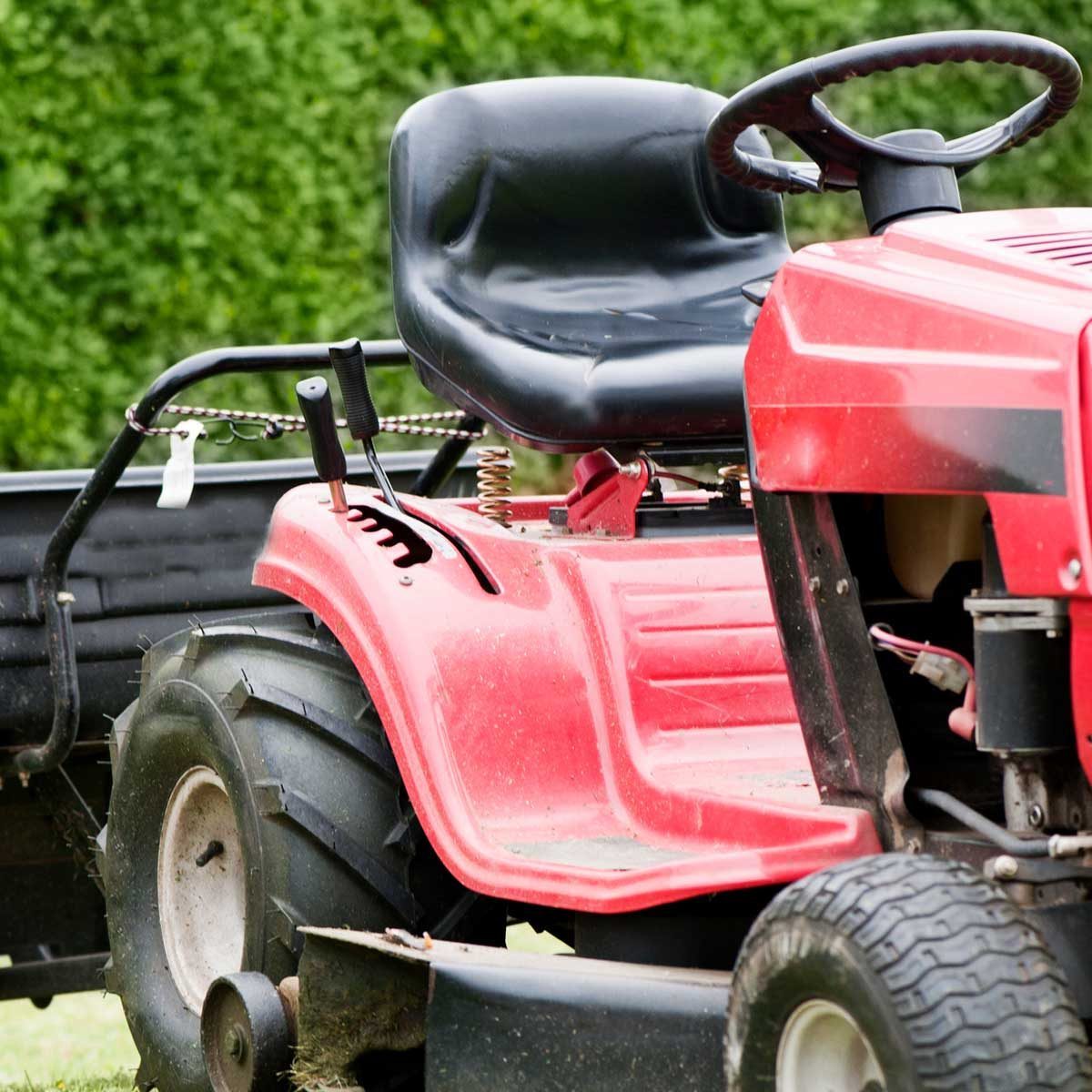 A red riding mower rests on grass, featuring a black seat and large rear tires, ready for lawn maintenance or landscaping tasks.