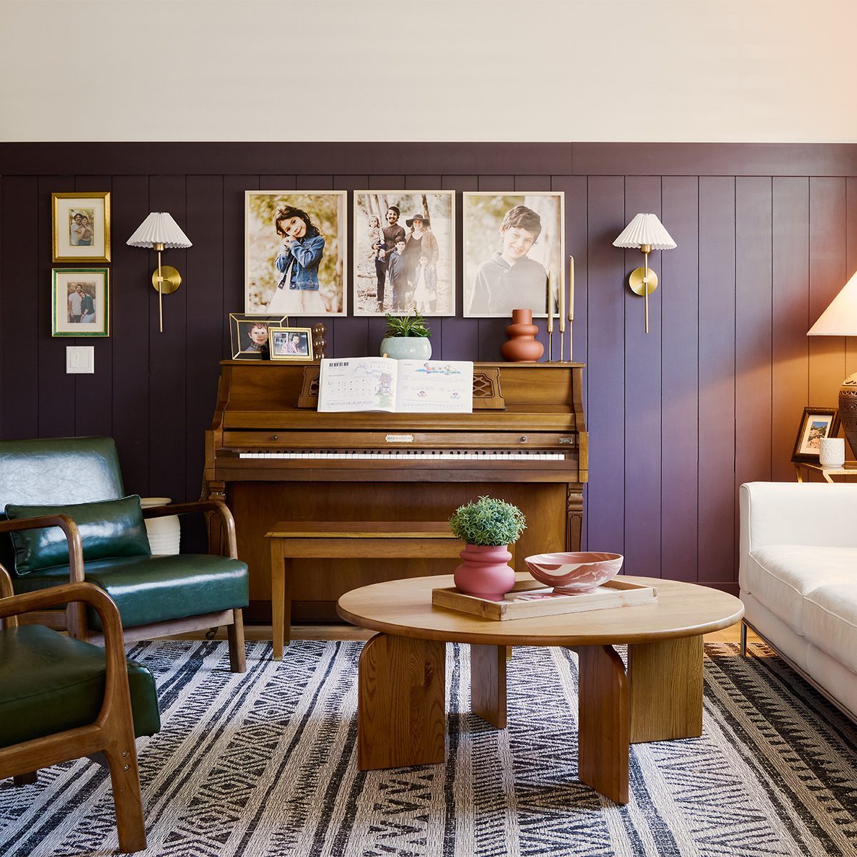 A cozy living room with a wood-paneled wall features a piano with sheet music, family photos, and sconces above. Green and white armchairs sit around a round wooden coffee table