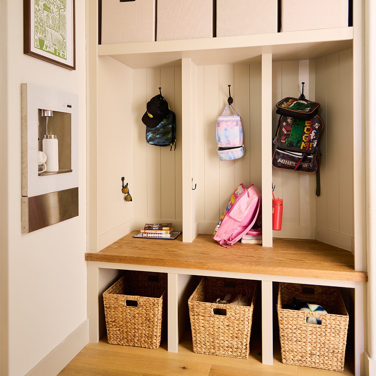 A neatly organized mudroom features three cubbies with hooks. Each cubby holds a backpack and some accessories. Below, there are three woven baskets for additional storage. The