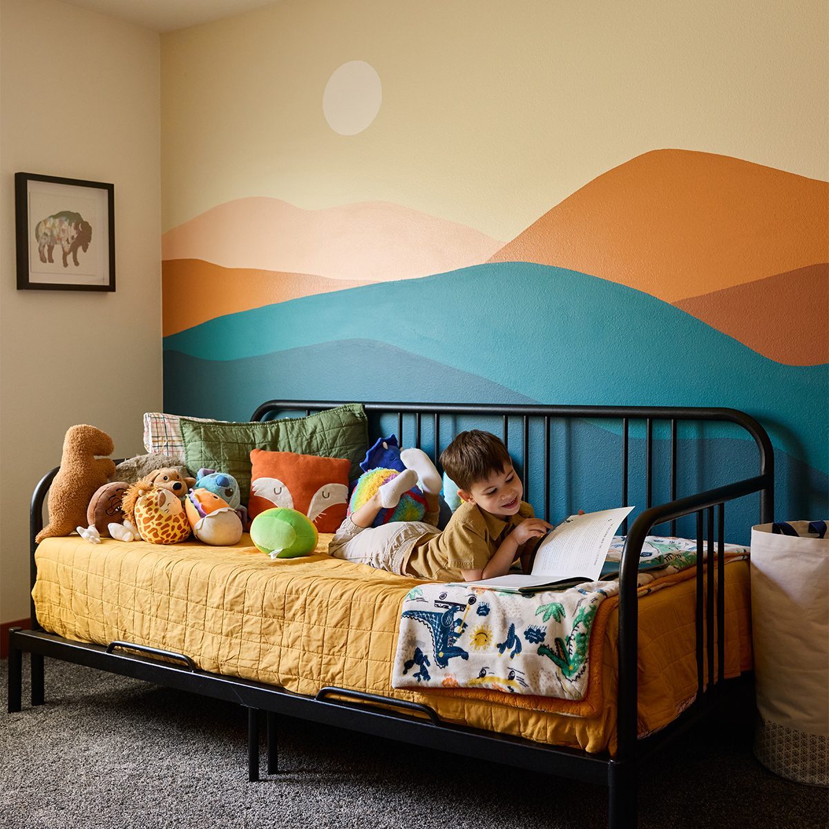 A young child lies on a bed with a yellow quilt, surrounded by plush toys. The wall behind the bed features a colorful mural of rolling hills in shades of blue, orange, and tan