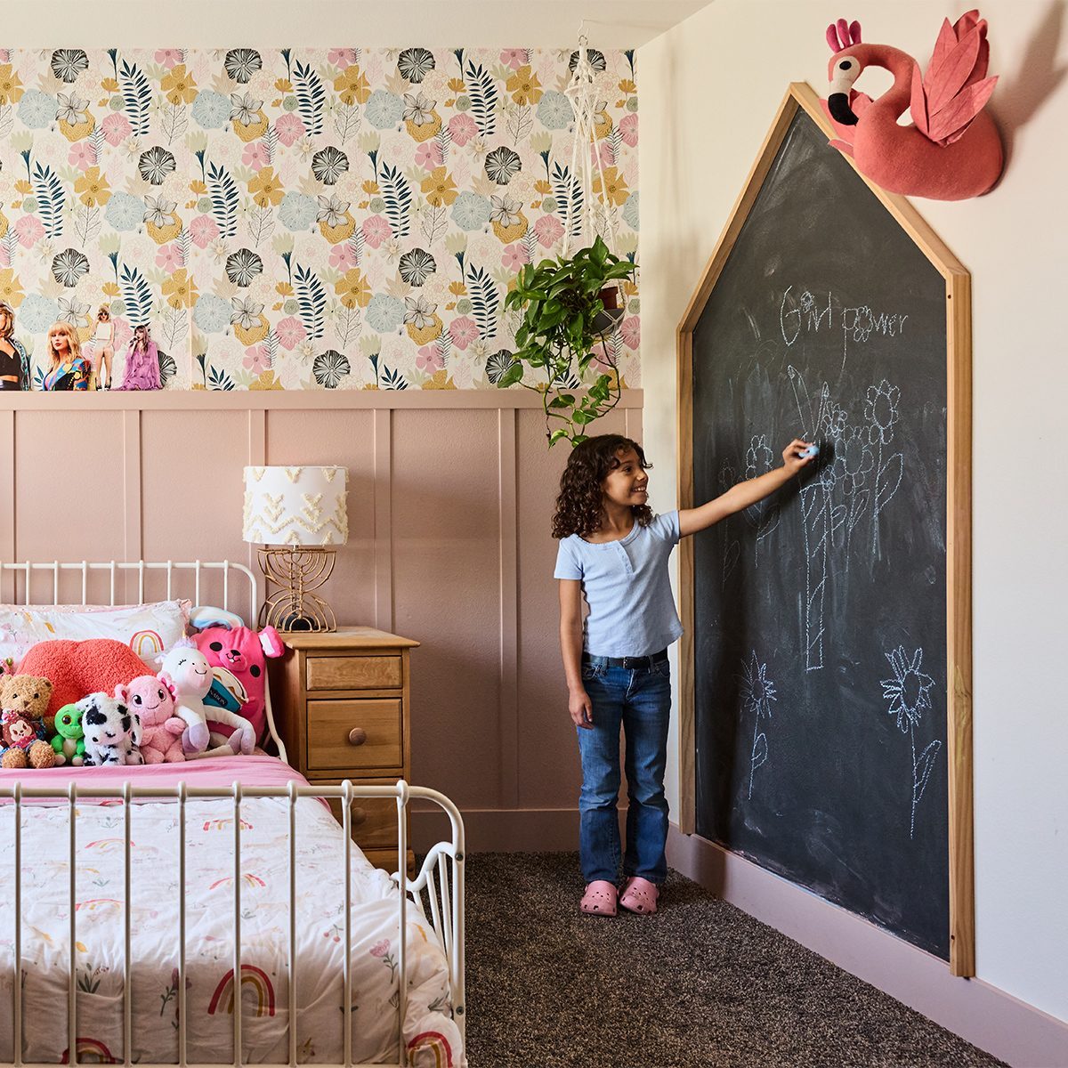A young child stands at a large chalkboard shaped like a house, drawing with chalk in a child's bedroom. The room features floral wallpaper, a bed with stuffed animals, a