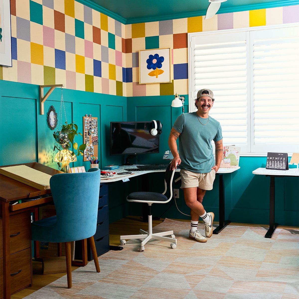 A person stands in a colorful, well-organized room featuring teal walls and a checkerboard pattern above. The room includes a desk with a computer, a drafting table, a chair with