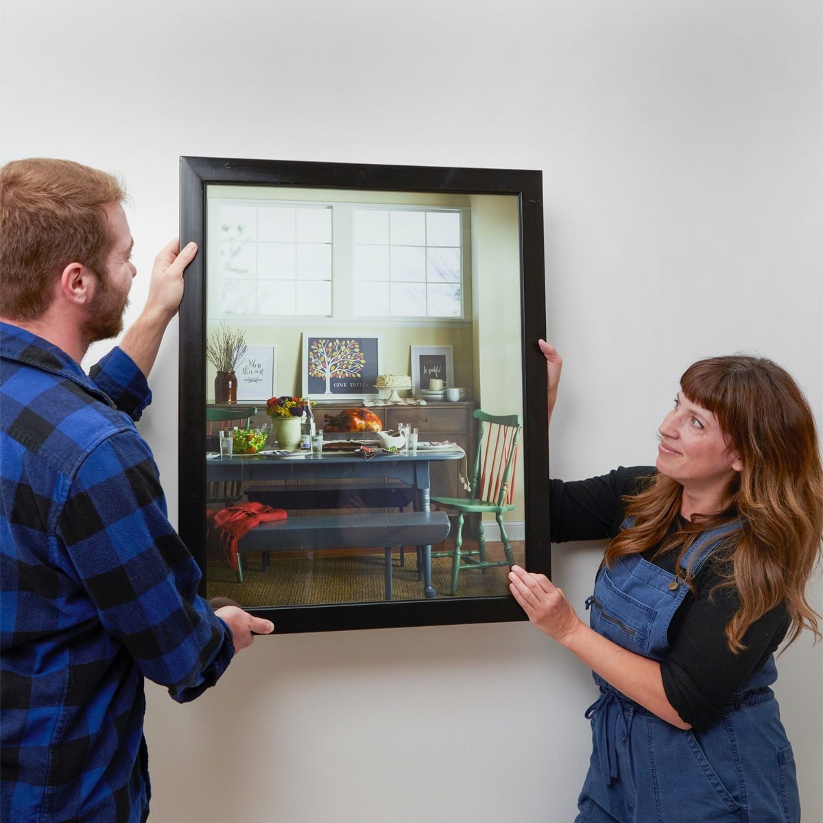 A man and woman hold a framed picture of a dining room, showcasing a table set with food and decorations, in a well-lit, cozy space.