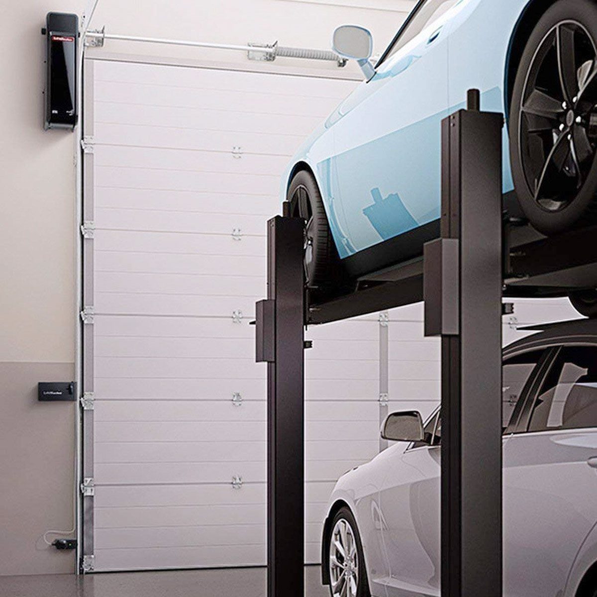 A blue car is elevated on a lift in a clean garage, with a white garage door and a gray car parked below it.