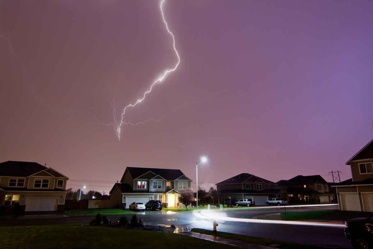 Lightning strikes the sky above a quiet neighborhood, illuminating houses and streets under a stormy, purple-hued night. Cars and a streetli