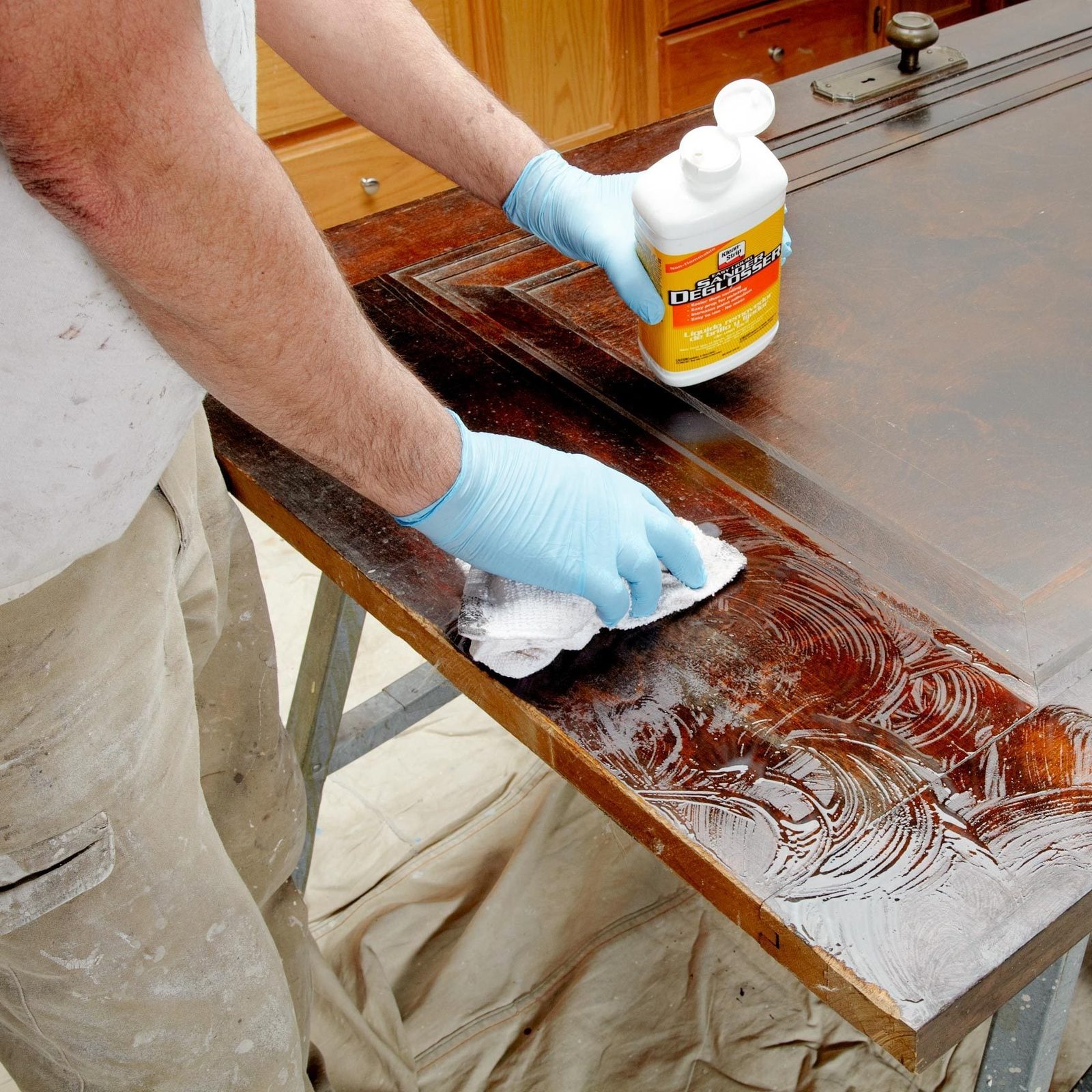 A person in blue gloves applies a cleaner to a wooden surface while wiping it with a cloth, in a workspace with wooden furniture and a drop