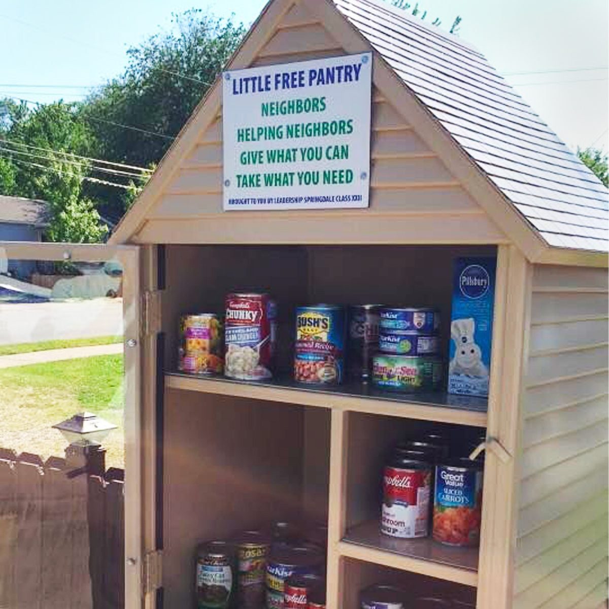 A small wooden pantry sits in a yard, offering various canned goods. It encourages community support with signs inviting neighbors to give a