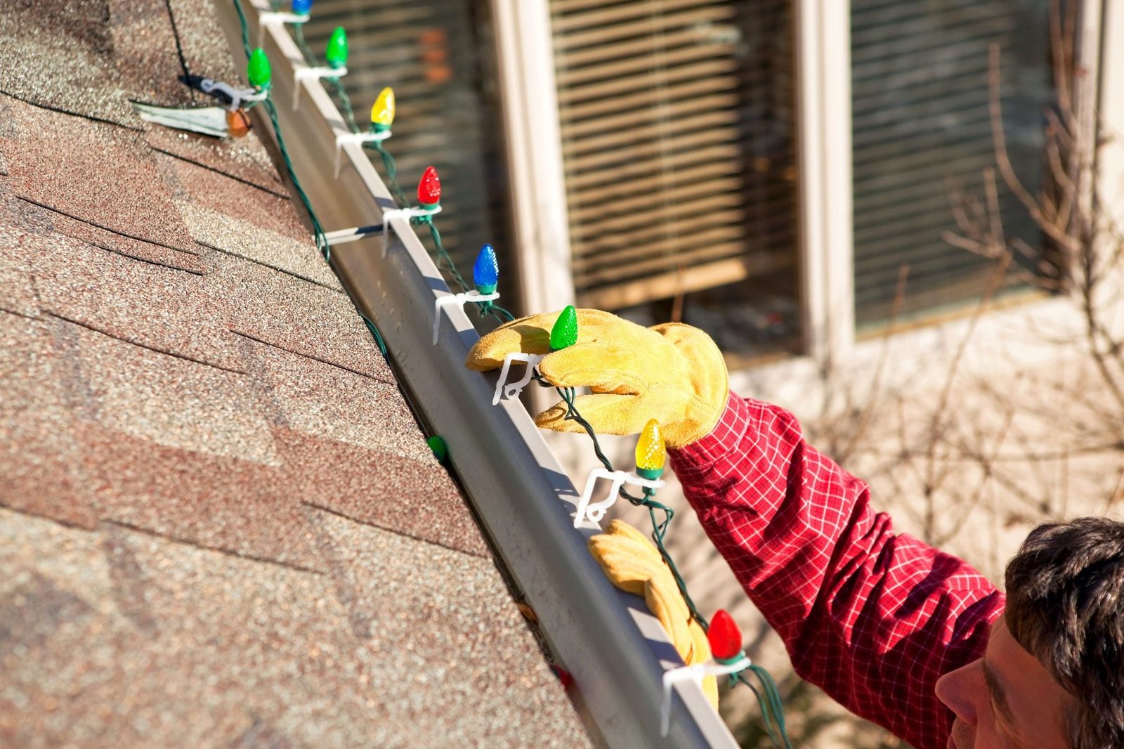 A person wearing gloves attaches colorful lights to a roof gutter on a sunny day, with a house and window in the background.