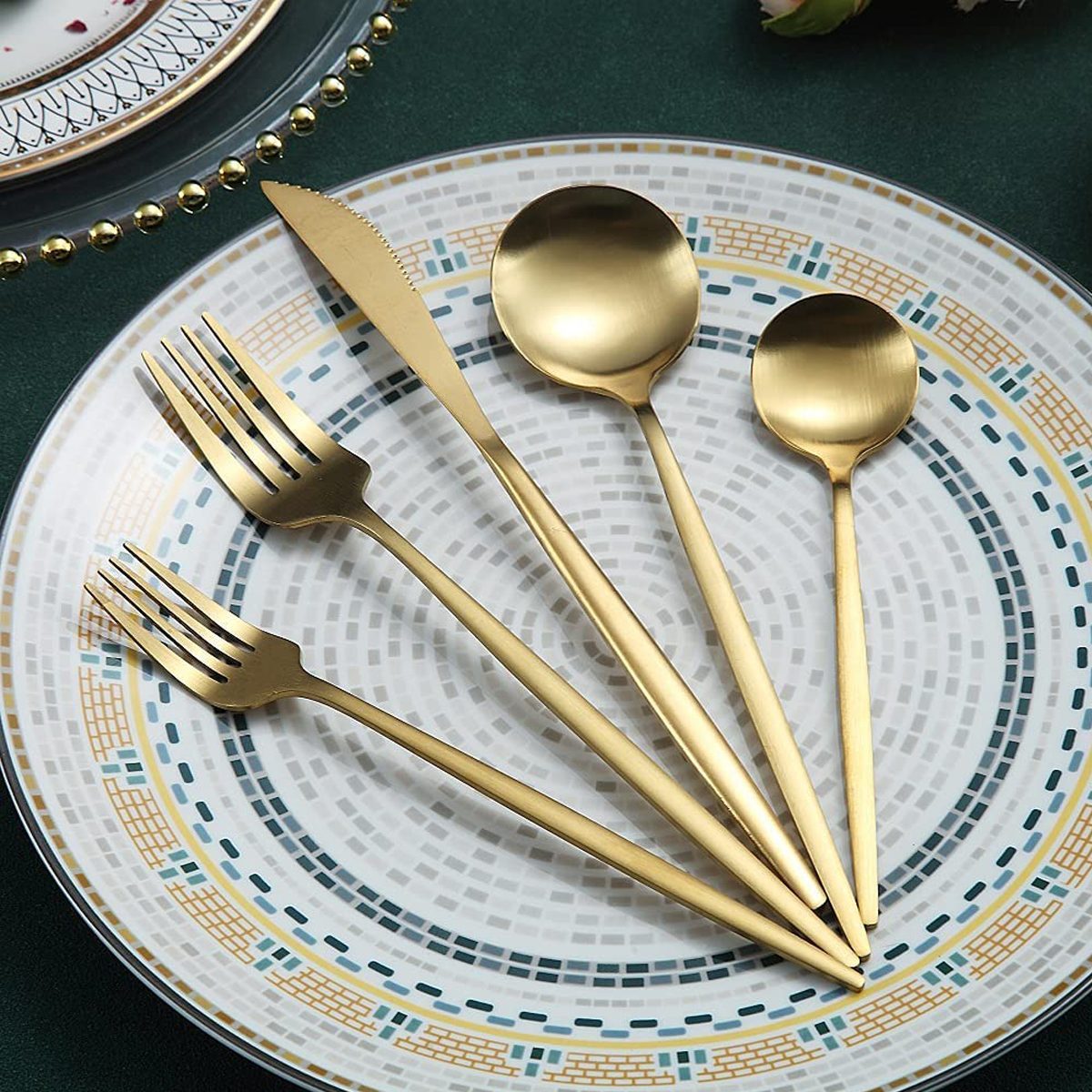 A set of gold utensils—two forks, a knife, and two spoons—rests on a decorative plate against a green background.
