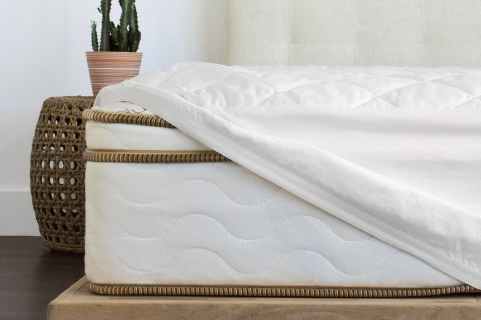A white mattress with a quilted top lies partially uncovered on a wooden base, beside a woven stool and a potted cactus in a bright room.