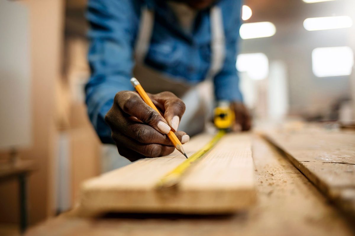 A hand holds a pencil, marking measurements on a wooden board. A tape measure is nearby in a workshop filled with soft light and wood shavin