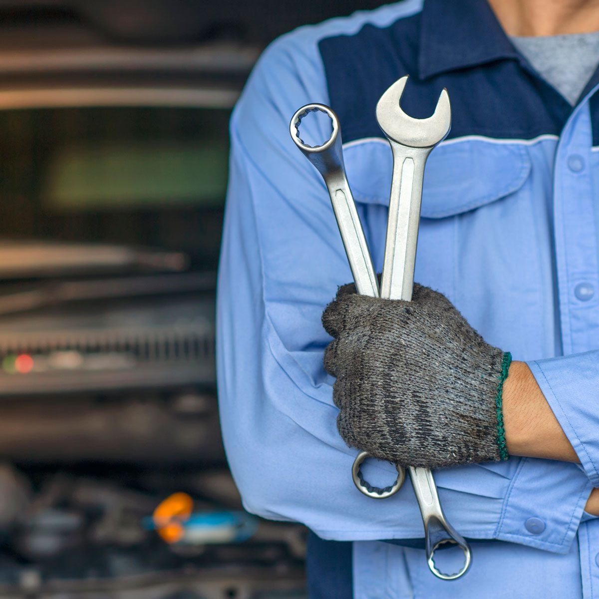 A person holds two wrenches in a gloved hand while standing in front of an open vehicle hood in a workshop setting.