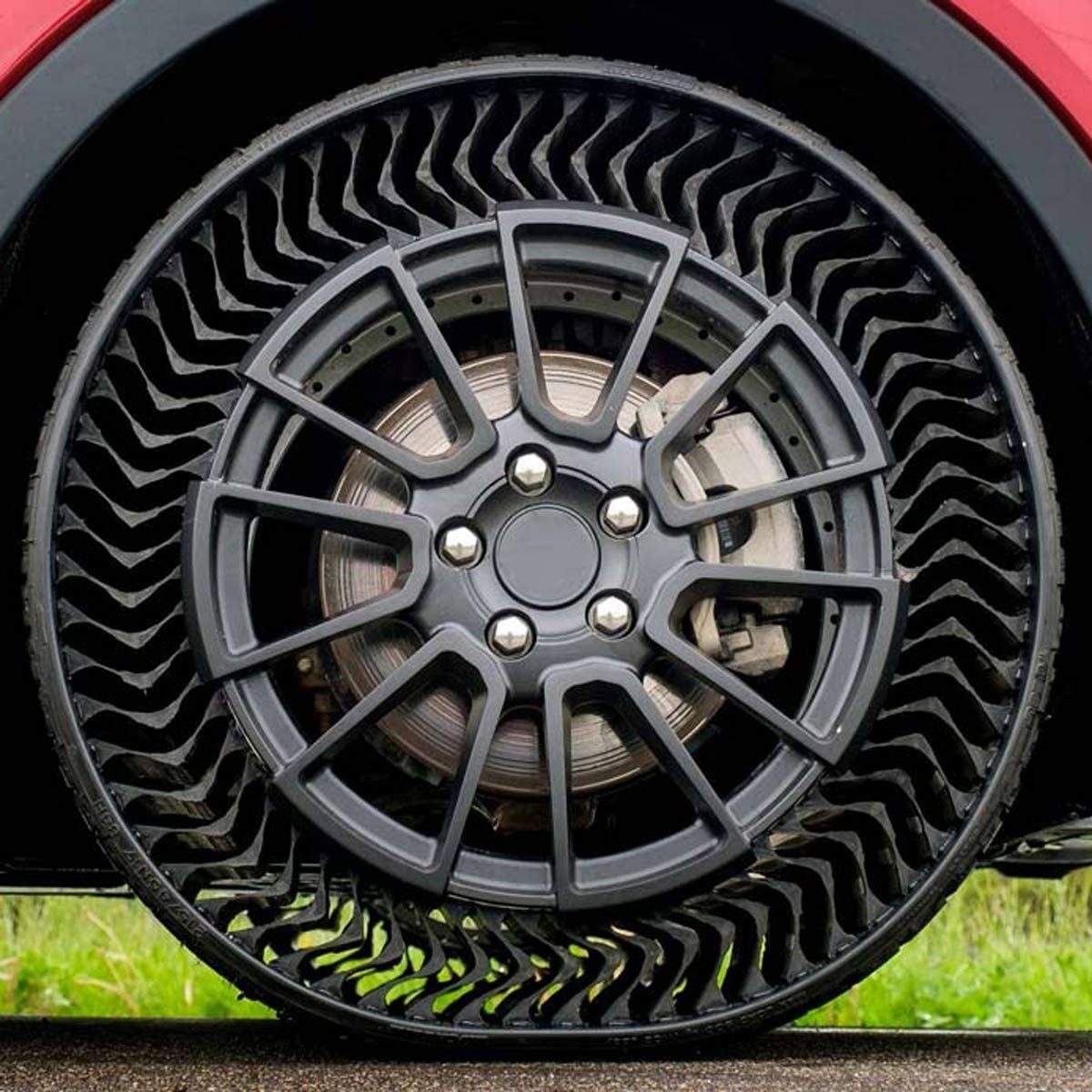 A unique tire with a honeycomb design is mounted on a car wheel, resting on a paved surface with greenery in the background.