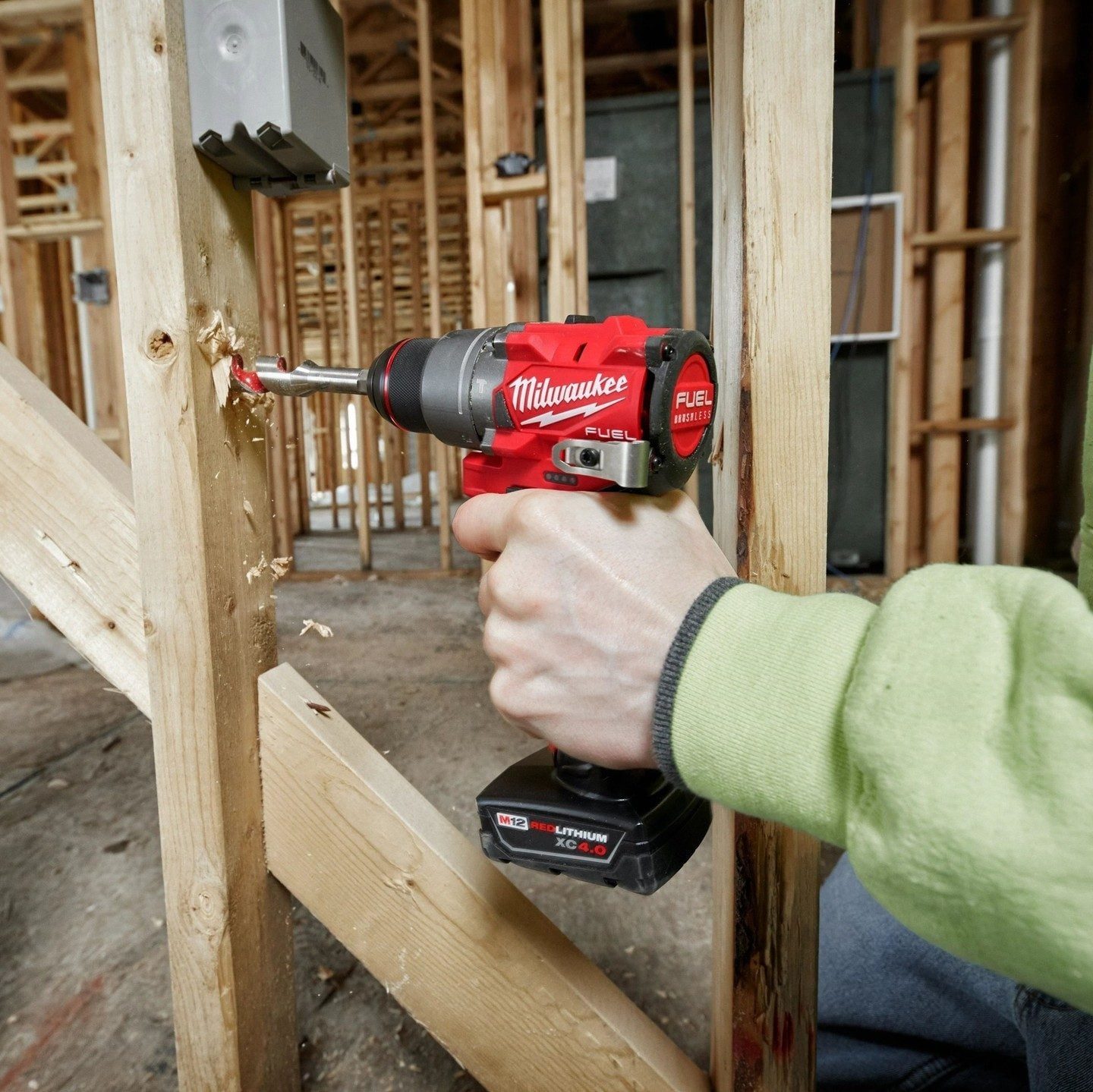 A person uses a red cordless drill to bore a hole into a wooden stud within a construction site. Surrounding structures are partially framed