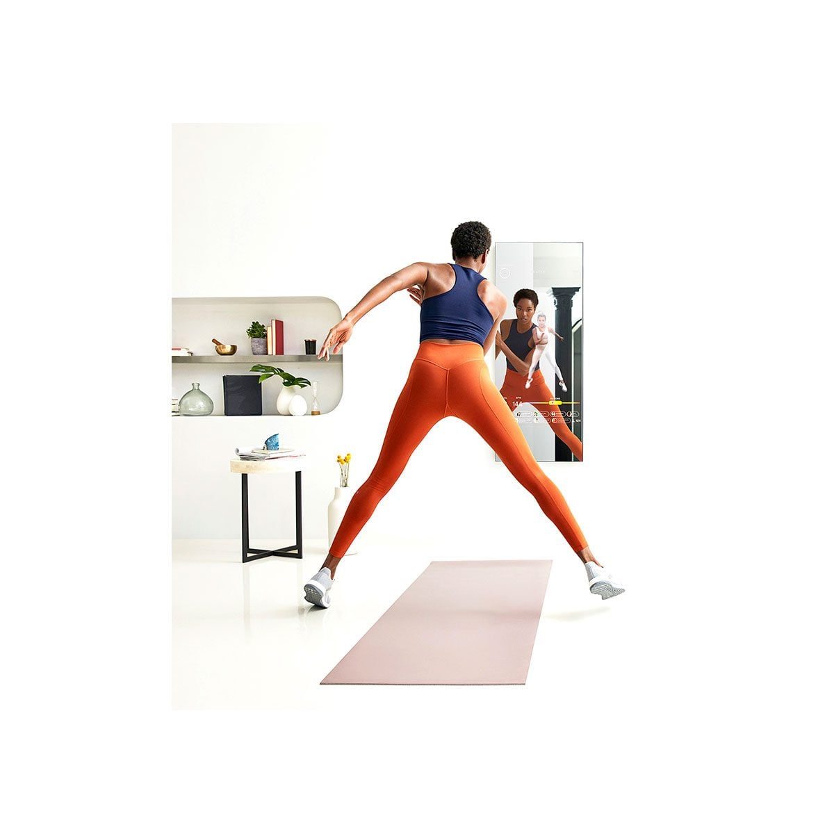 A person in orange leggings exercises on a mat while observing their reflection in a mirror, surrounded by a minimalist room with decor item