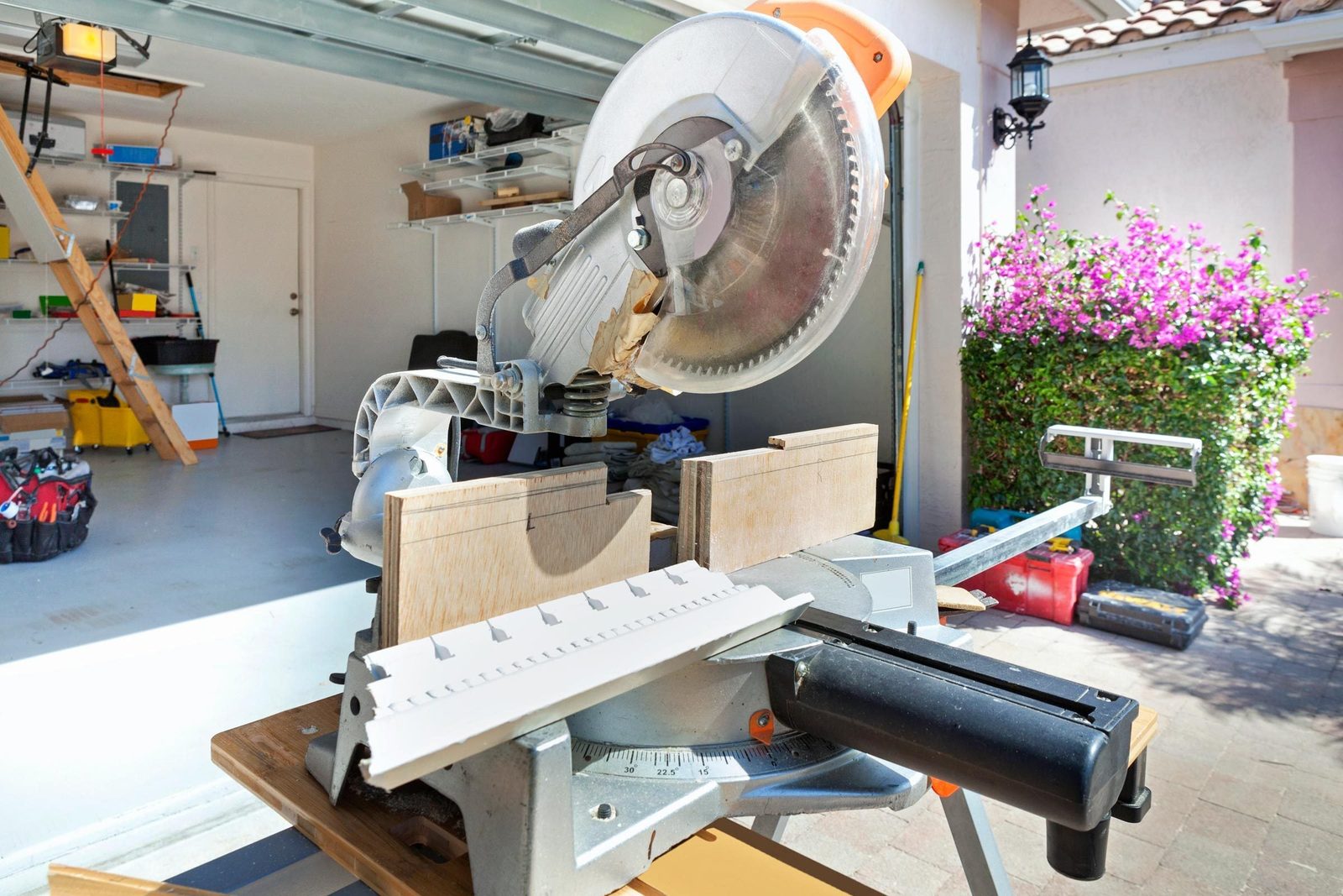 A miter saw rests on a workbench, ready to cut wood, surrounded by a garage space with shelves and blooming flowers outside.