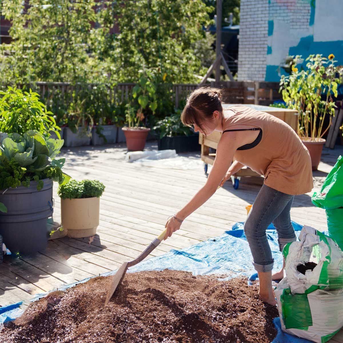 A woman shovels soil on a blue tarp in a garden, surrounded by potted plants, while sunlight illuminates the wooden deck.