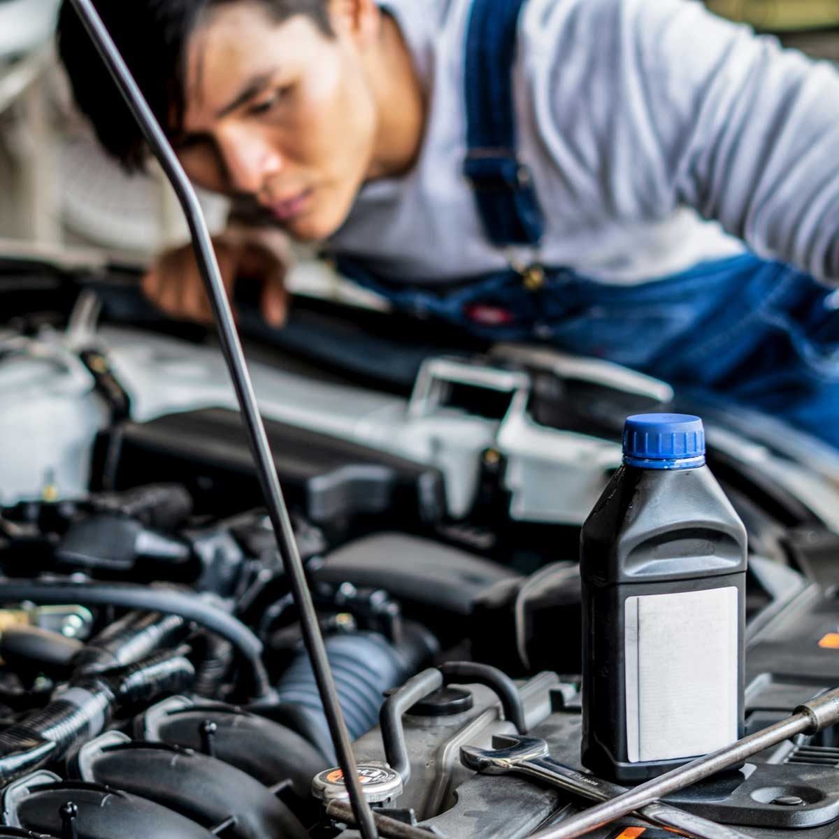 A bottle of oil rests on a car engine while a person leans in closely, inspecting the engine components in a garage setting.