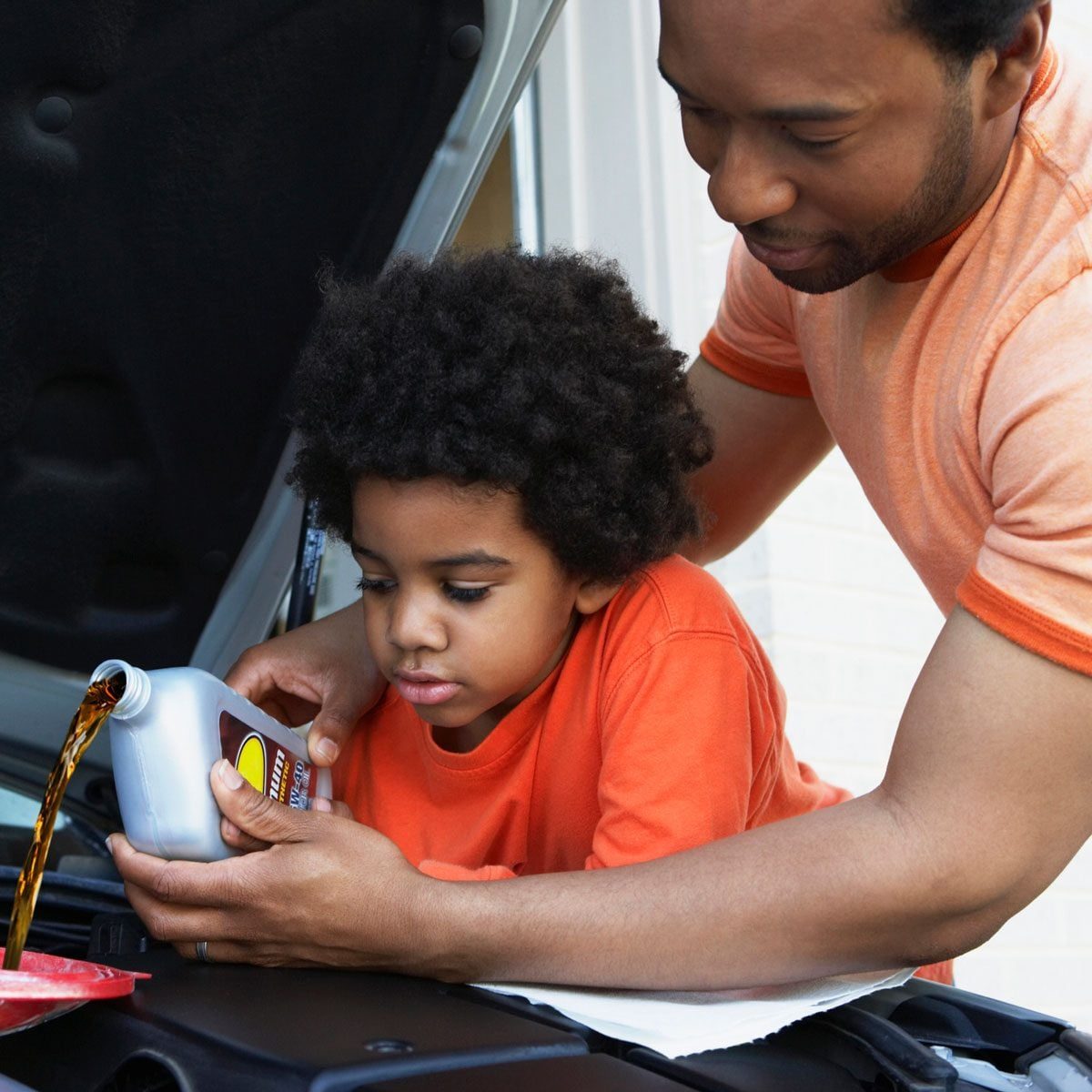 A father assists his young son in pouring motor oil into a car's engine while they both lean over the open hood of the vehicle.