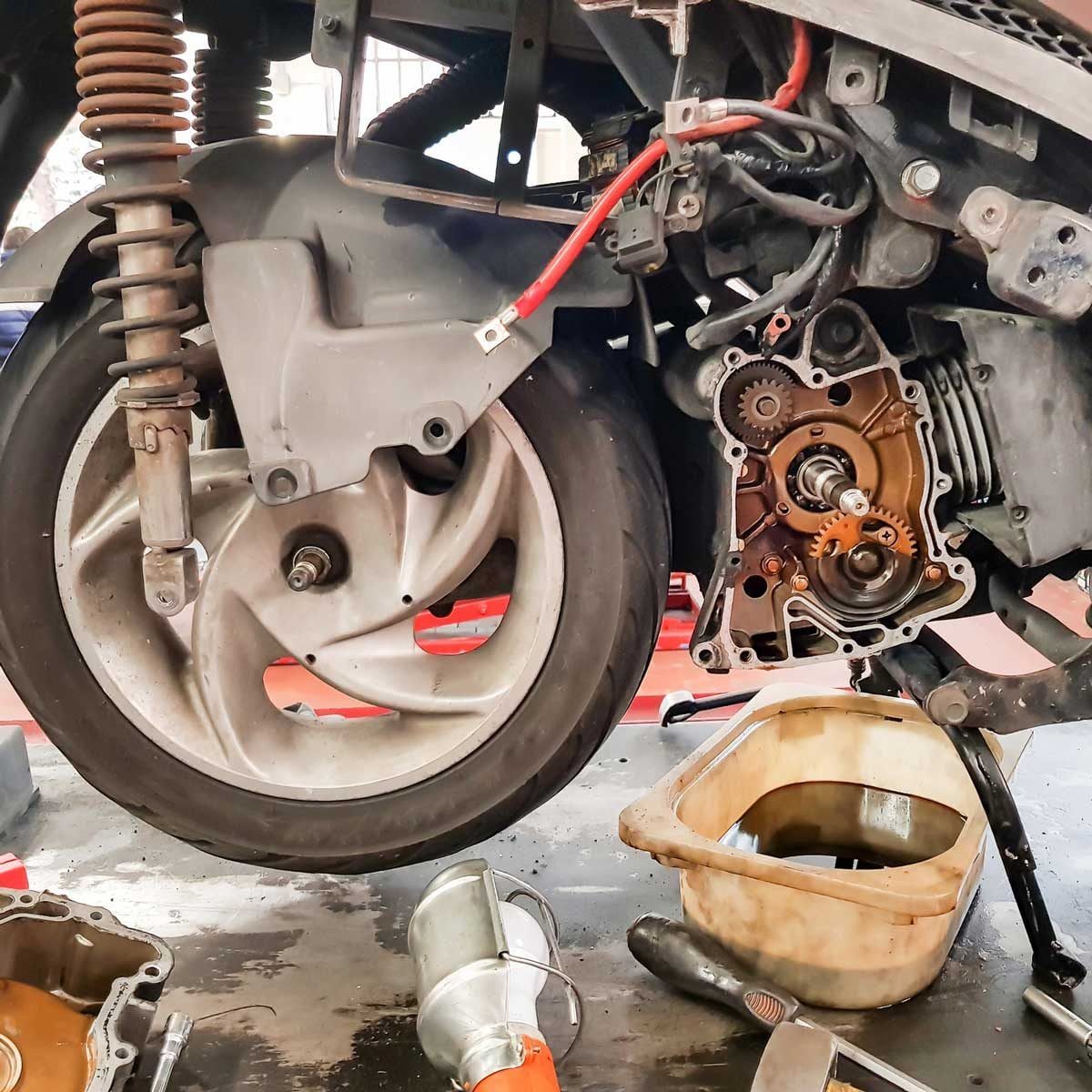 A scooter's rear wheel is disassembled, revealing internal gears and components, with tools and an oil catch pan scattered on the workshop f