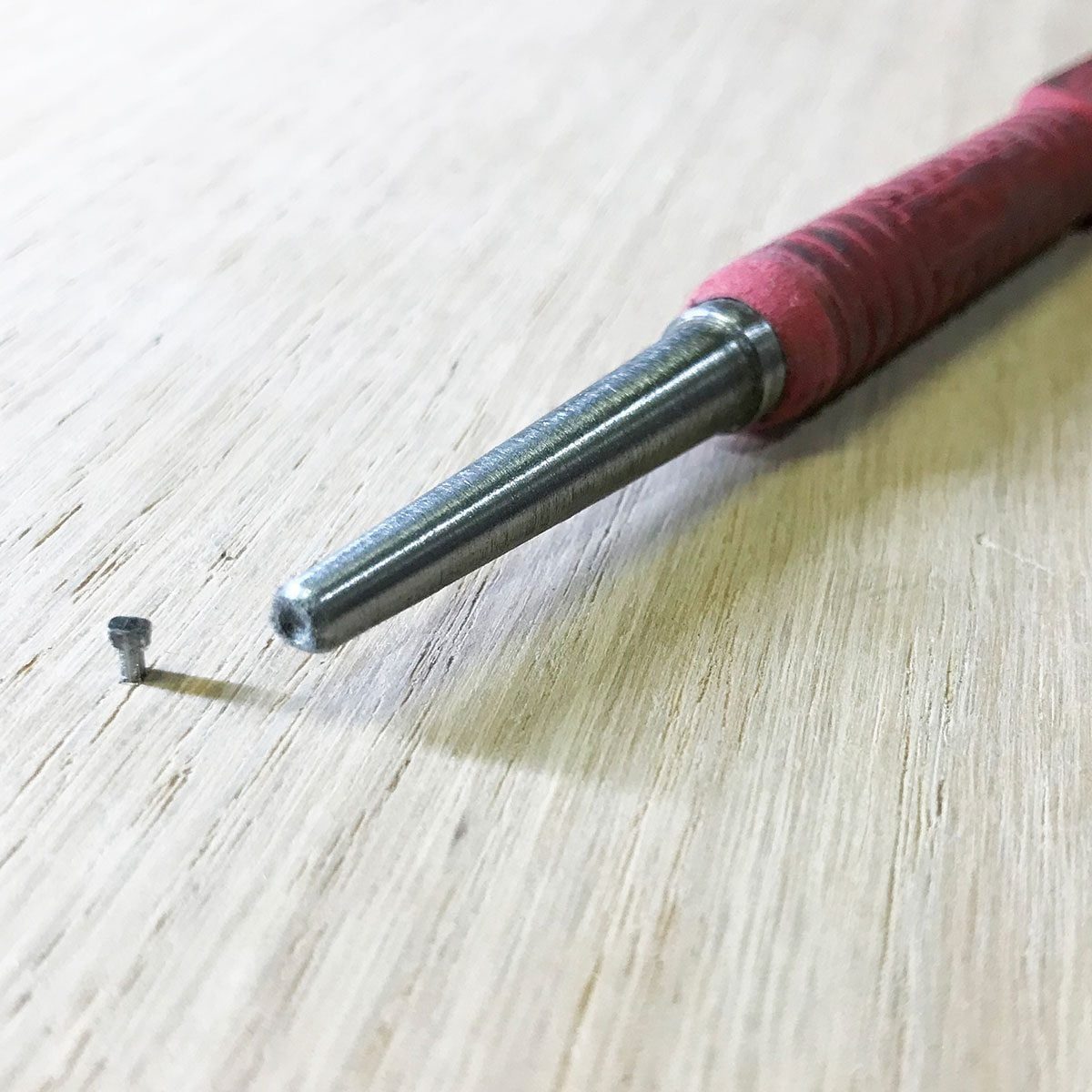 A pointed metal tool rests near a small screw on a smooth wooden surface, suggesting a task related to assembly or repair.