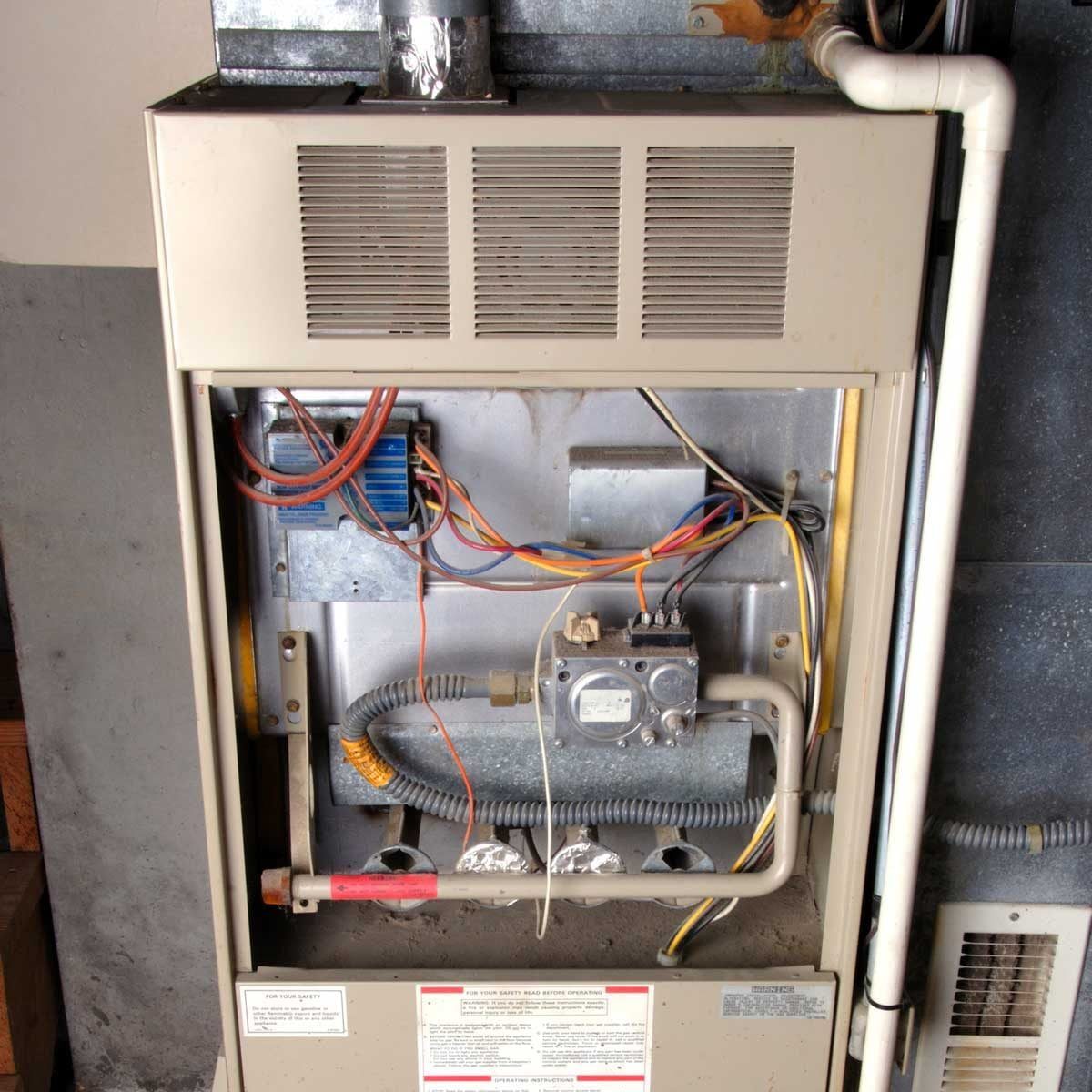A furnace unit is displayed, showing exposed wires and components, while it operates in a dimly lit utility area with a concrete wall.