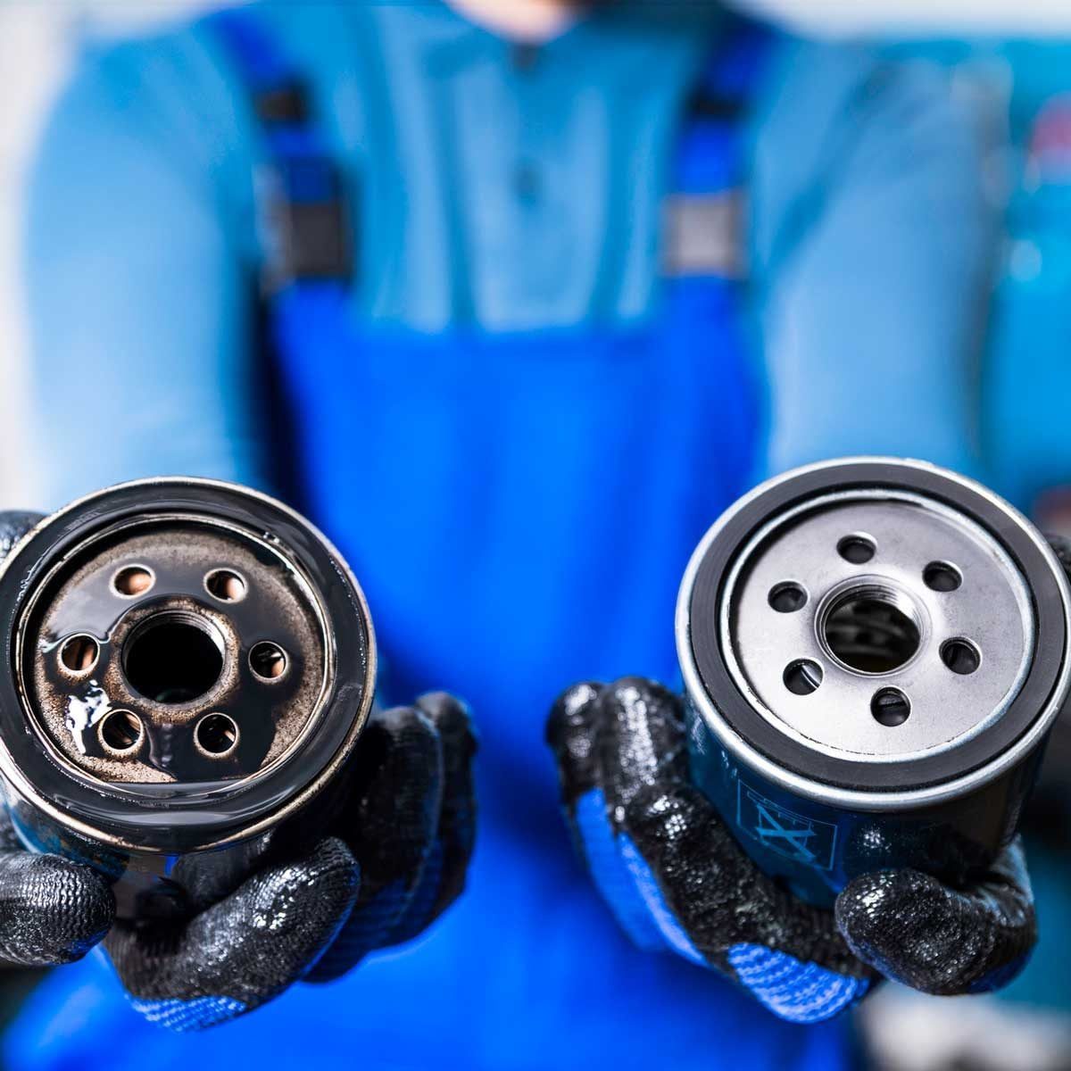 A person holds two oil filters, one dirty and one clean, demonstrating the difference while wearing blue gloves in a workshop setting.