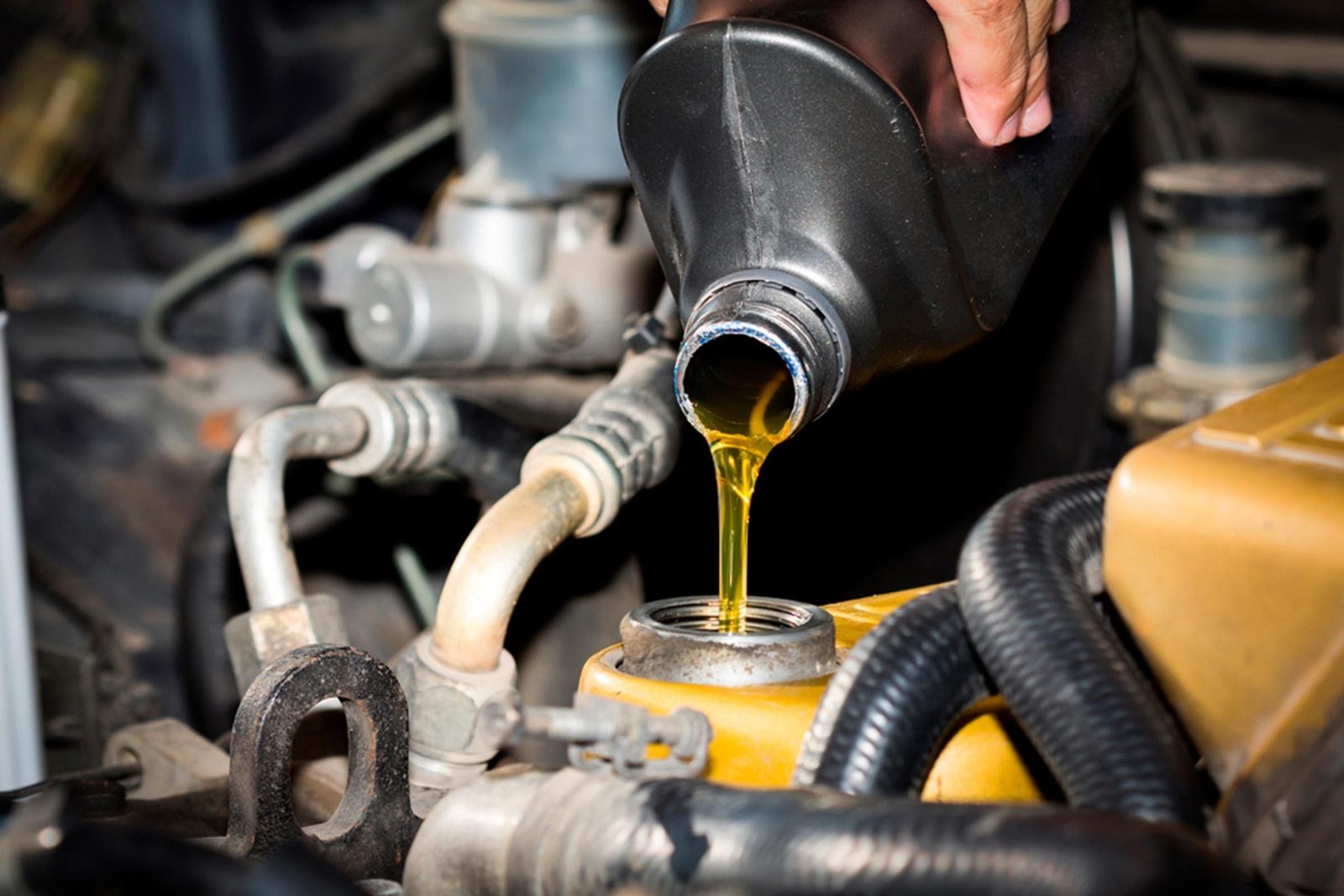 A hand pours yellow liquid from a bottle into an engine component surrounded by various hoses and machinery within a vehicle's engine bay.
