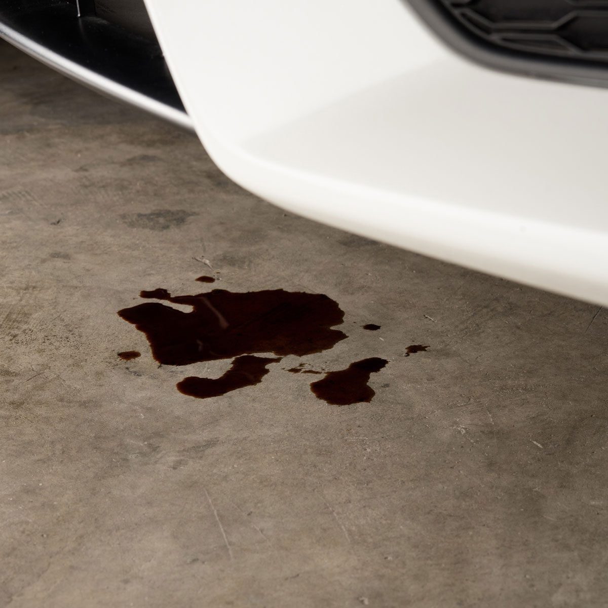 A white car is parked, leaking a dark brown fluid onto a concrete floor, creating a splash mark beneath the vehicle.
